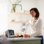 Frau giesst Eierlikoer aus Vitamix Aer Disc Container in Tasse, Ascent Mixer im Hintergrund