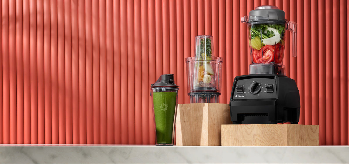 Vitamix Explorian E310 con Personal Cup Adapter e smoothie verde davanti a una parete a lamelle rosse