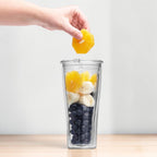 Hand fuellt Orange in Vitamix To-Go Cup mit Bananen und Blaubeeren auf Holzarbeitsflaeche
