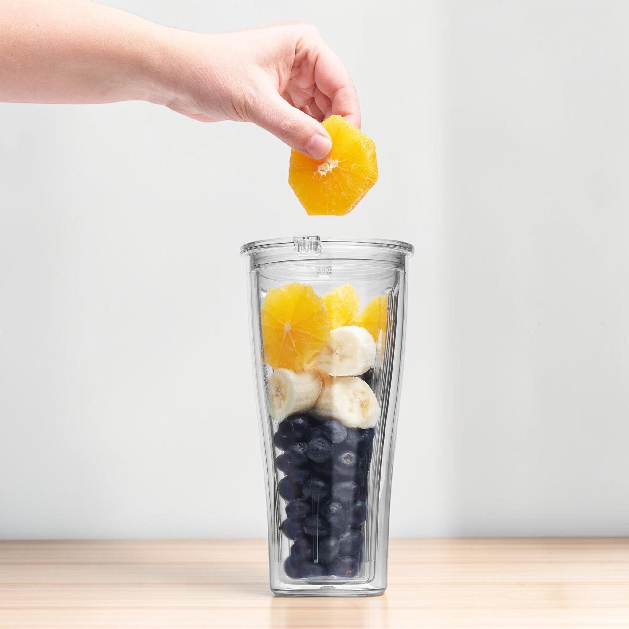 Hand fuellt Orange in Vitamix To-Go Cup mit Bananen und Blaubeeren auf Holzarbeitsflaeche